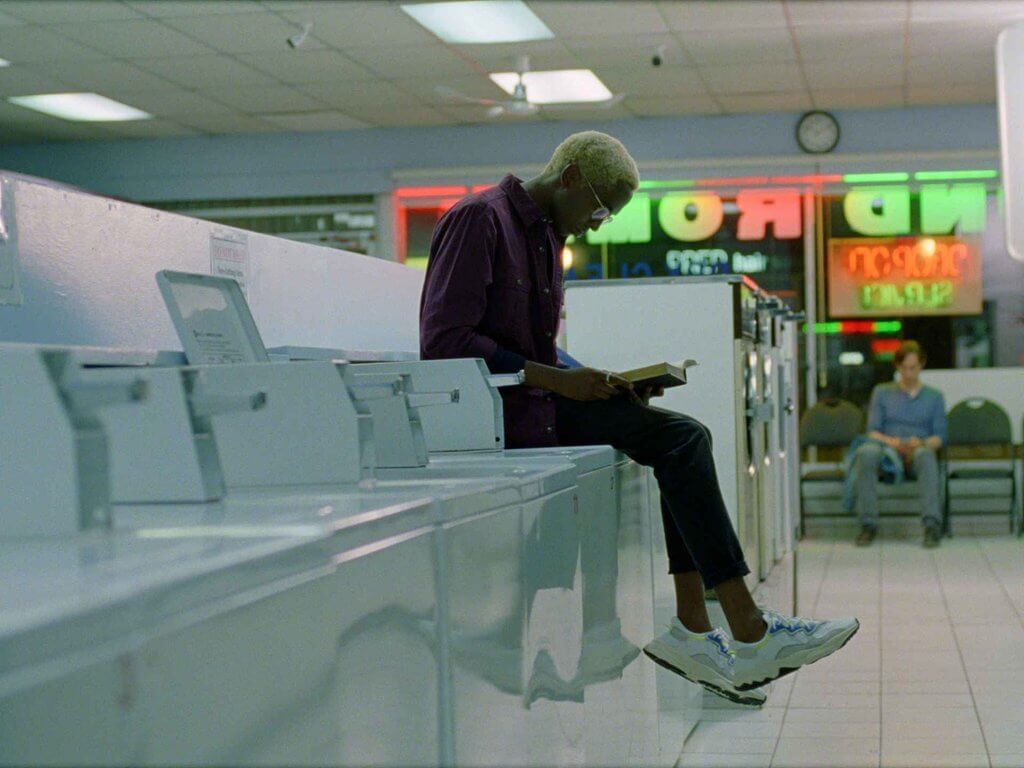 A man with glasses sitting on a washing machine in a laundromat