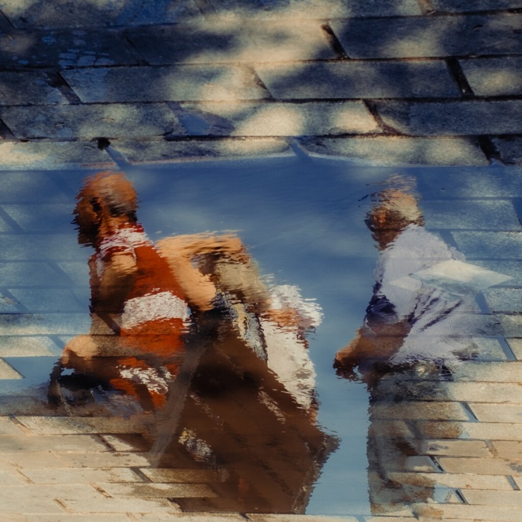 The Distorted Reflection Of People In A Pool Of Water On A Brick Floor, Creating An Intriguing Mix Of Textures And Shapes