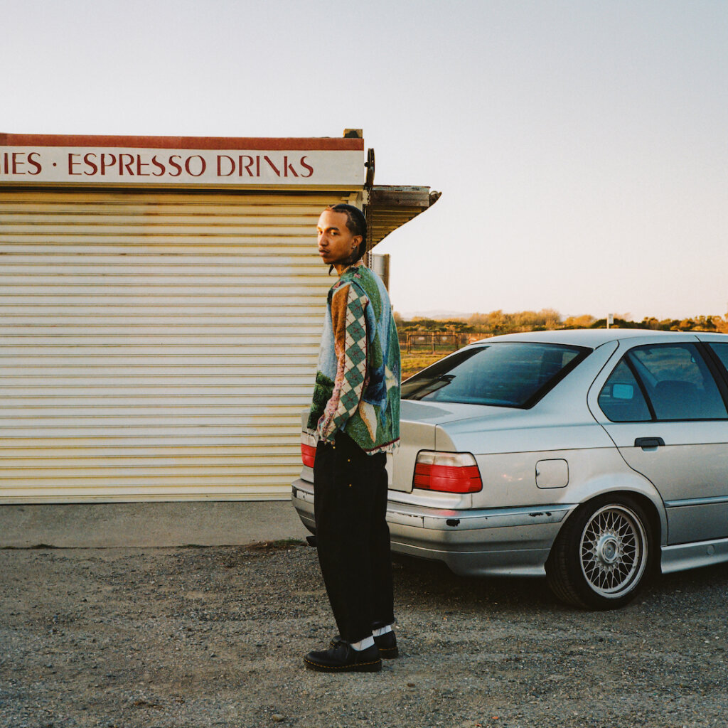 A Man Stands Near His Silver Car Wearing Black Pants And A Green Sweater