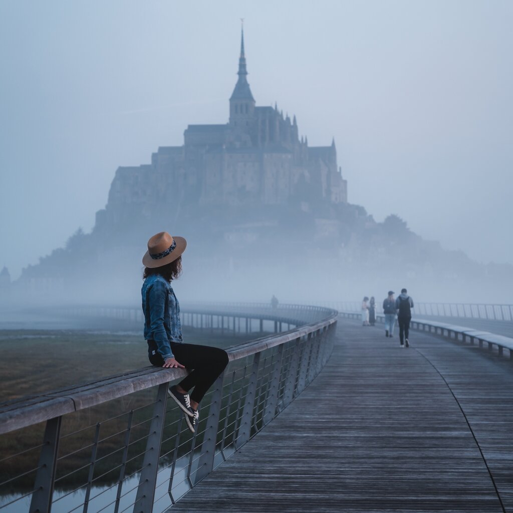 A Woman Wearing A Hat Sits On A Bridge Ledge While Others Walk Towards A Foggy Castle