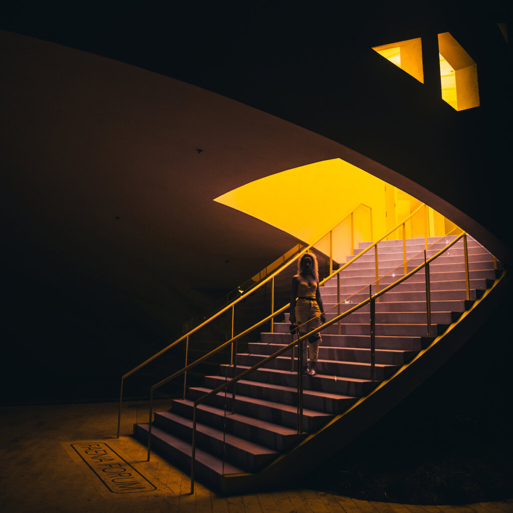 A Woman In A Yellow Dress Stands On A Staircase With Yellow Handrails In A Brightly Lit Architectural Building With Reflective Windows