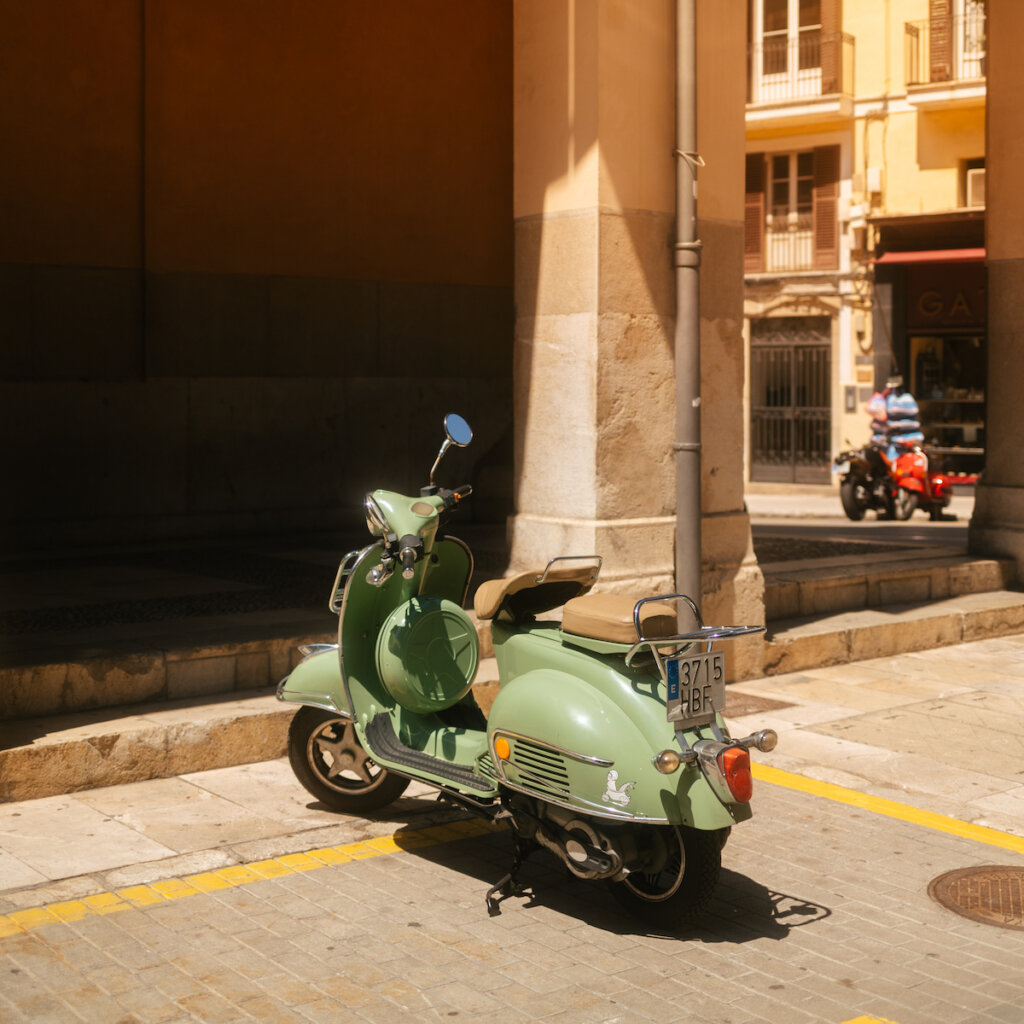 An Old Green Motorcycle Parked Near A Building With Arches In A City