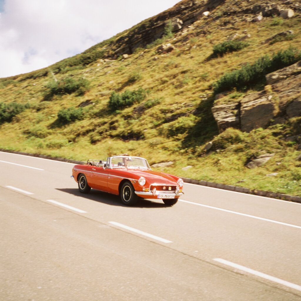 A Vintage Red Convertible Drives On A Highway Next To A Grassy Mountain