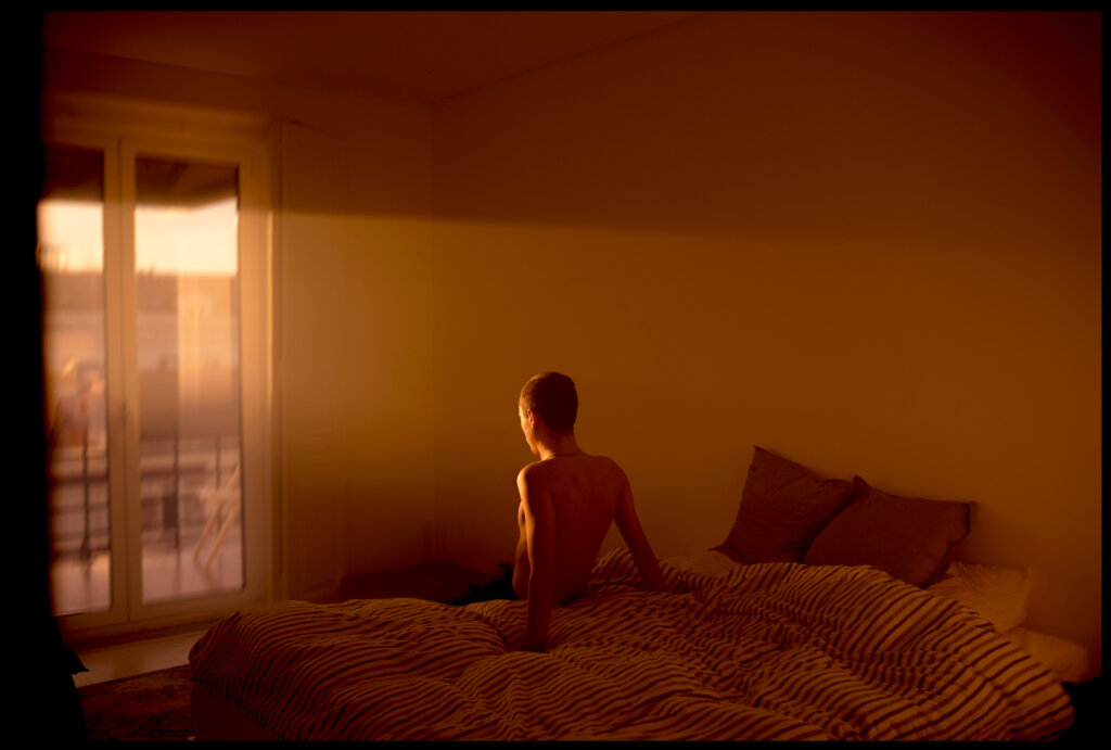 A Man Sitting On A Bed In His Bedroom Looking At The Sunrise Through The Balcony Doors