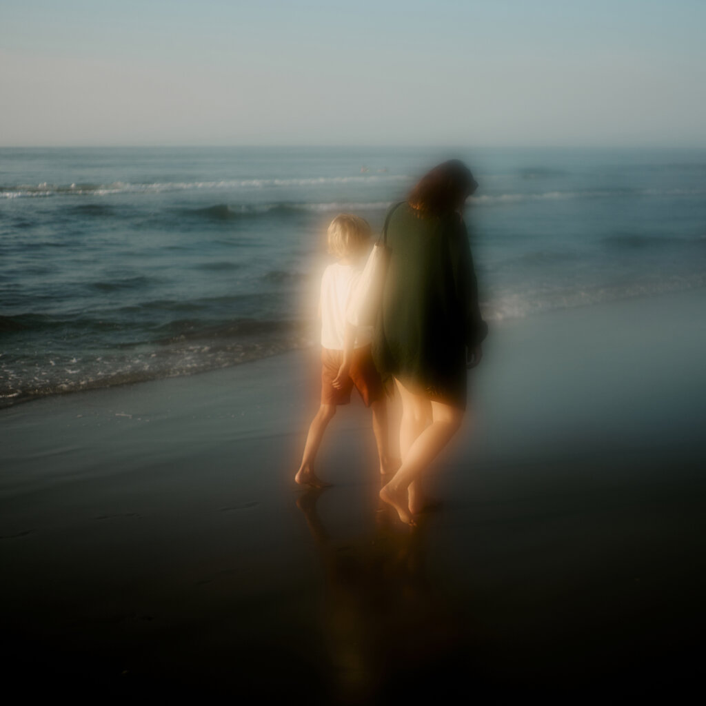 A Woman Wearing Green And A Boy Are Walking Barefoot On The Beach At Dawn