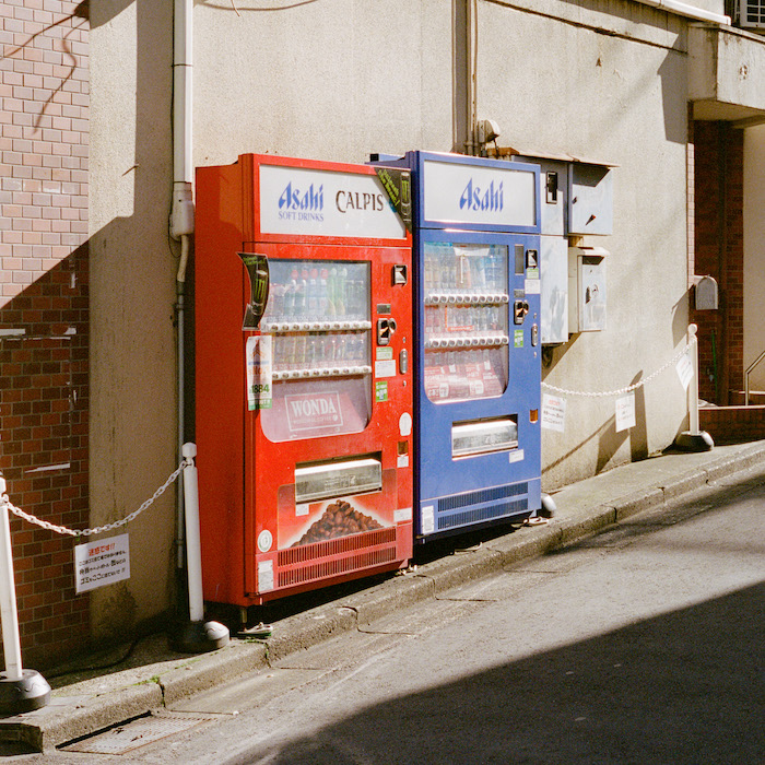 Two Beverage Vending Machines, One Red And One Blue, Positioned Side-by-side On A Sidewalk Next To A Building