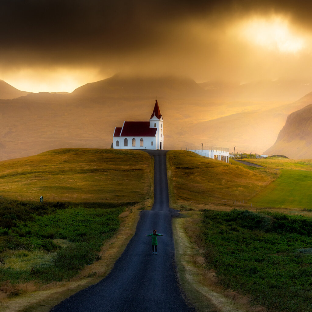 A Person Stands On A Road With Spread Arms In Front Of A White And Red Church Sitting On A Hill