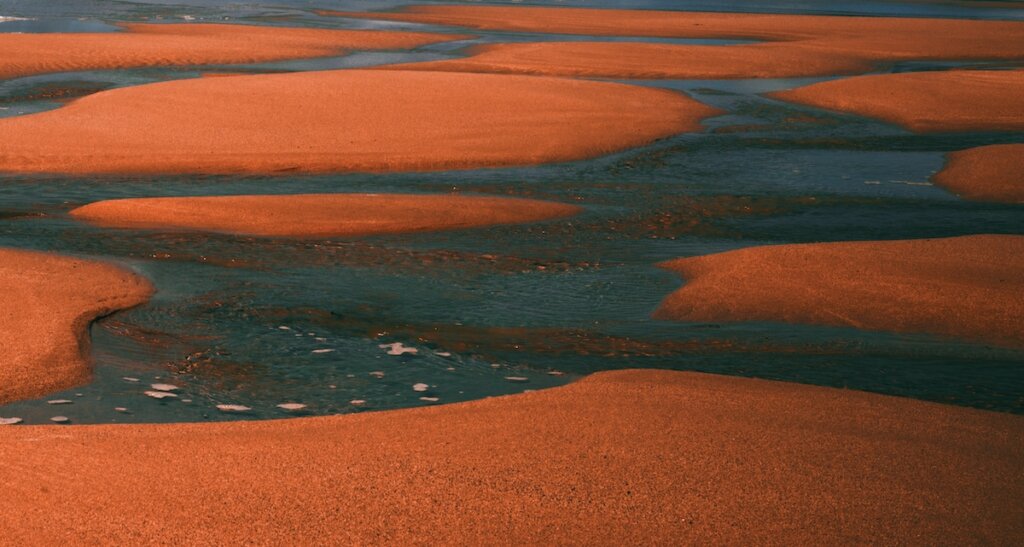 A River Winds Through Orange Sand Creating A Path Of Varying Widths And Curves