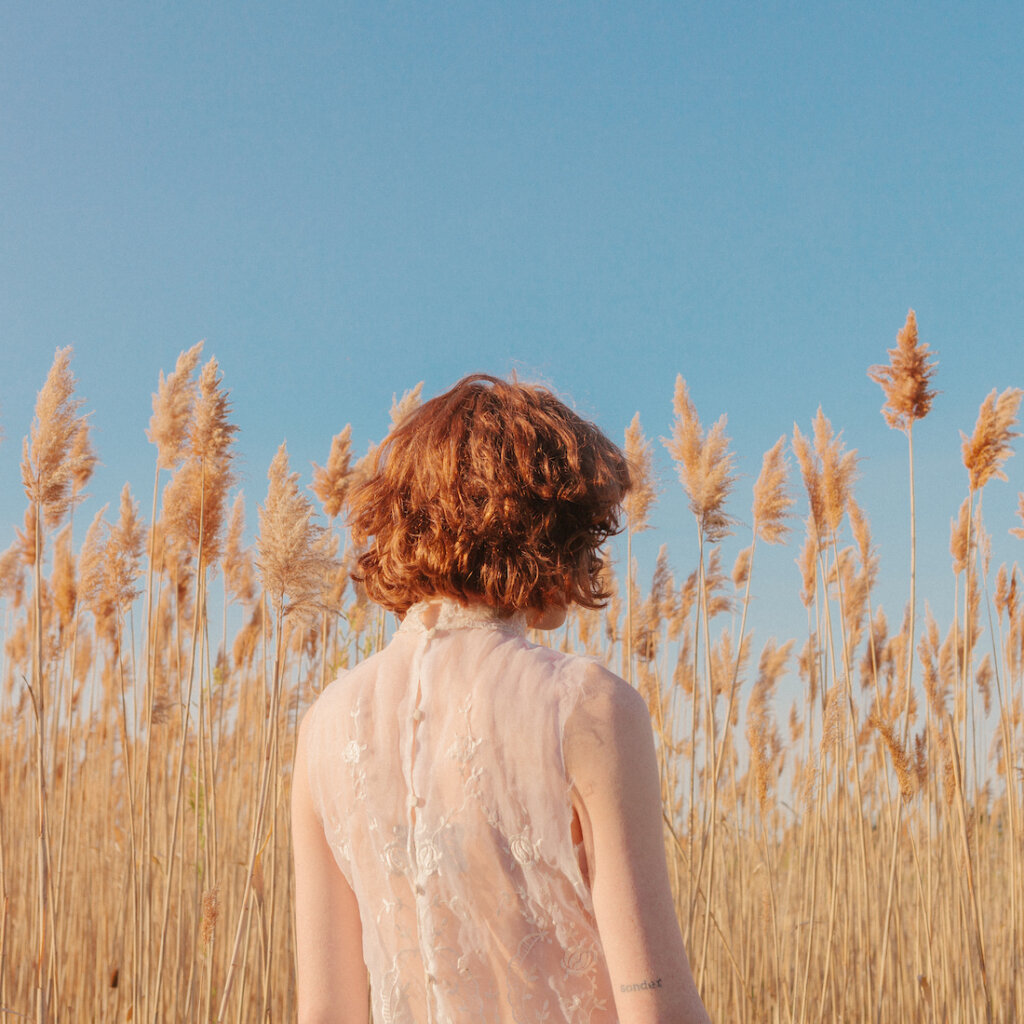 A Woman With Vibrant Red Hair Stands In A Field, Wearing A White Transparent Blouse, Surrounded By The Tall Phragmites Australis Reeds That Sway Gently In The Breeze