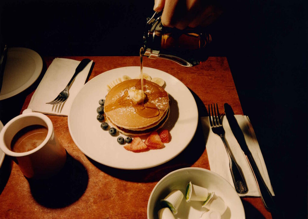 In A Dimly Lit Diner, A Hand Pours Syrup Onto A Pancake Stack Surrounded By Banana Slices, Strawberries, And Blueberries, With Butter, Mug, Cutlery.