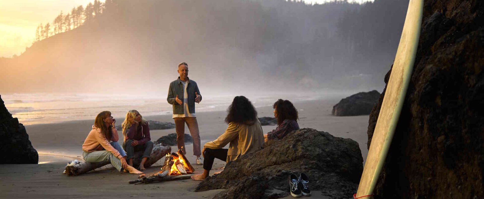small group gathered around a beach bonfire at dusk, with a wetsuit and surfboard nearby
