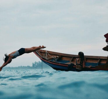 A Woman In A Striped Swimsuit Dives Gracefully From A Wooden Boat